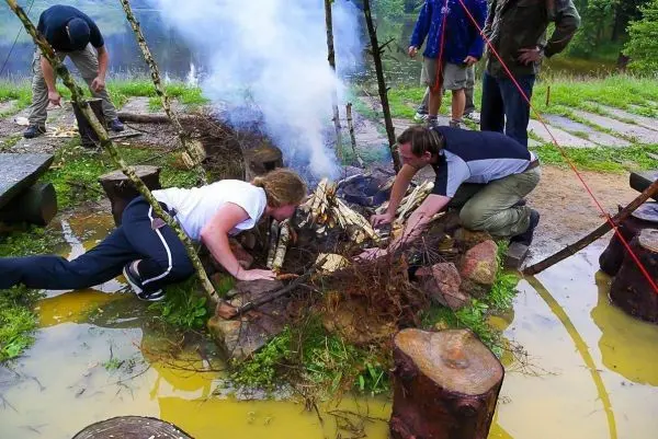 Kleine Flamme mit Rauch zwischen Holzstücken und zwei Personen, die Zunder hinzufügen