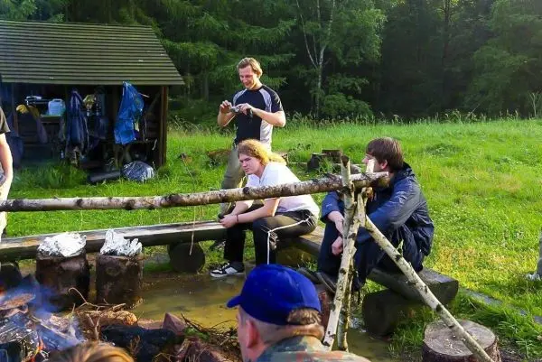 Person sitzt an Feuerstelle und beobachtet andere beim Kochen im Outdoorkurs