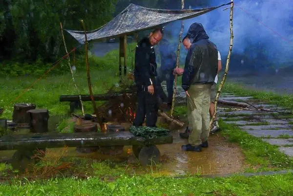 Tarp-Notunterkunft schützt vor Regen, während Holzscheite um ein kleines Bodenfeuer angeordnet sind