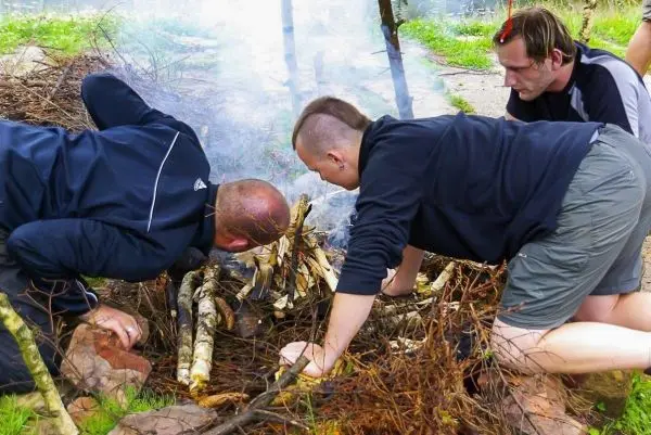 Zunder glimmt in einem kleinen Feuer, während drei Personen darüber gebeugt sind