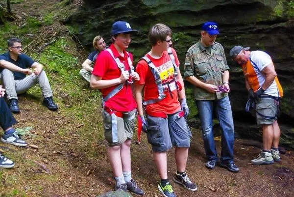 Zwei Jugendliche mit Klettergurten stehen in einem Wald, während andere Personen im Hintergrund sitzen