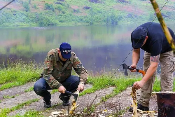 Zwei Personen bearbeiten Holz mit Äxten am Ufer eines Gewässers