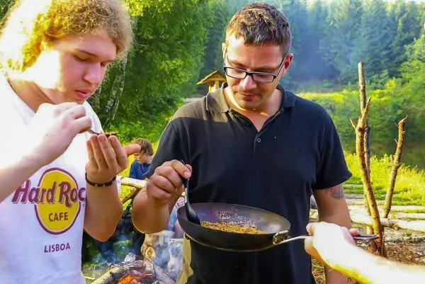 Zwei Personen betrachten gemeinsam eine Pfanne mit geröstetem Essen über einem Lagerfeuer