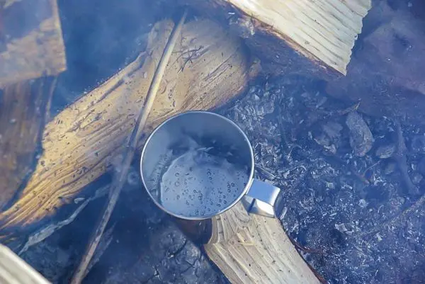 Edelstahlbecher mit Wasser über glühenden Holzscheiten erhitzt