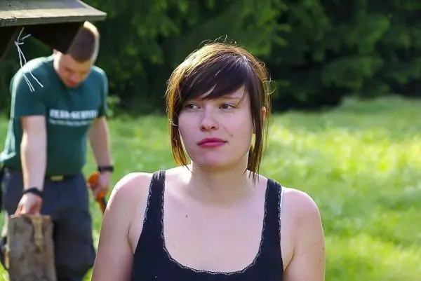 Frau mit kurzer Frisur steht im Vordergrund, im Hintergrund Holz wird bearbeitet