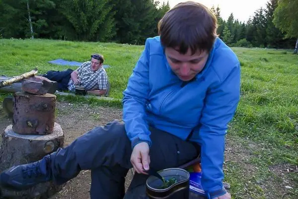 Frau rührt in einem Topf auf einem Holzstamm, Mann sitzt im Hintergrund