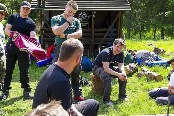 Gruppe sitzt auf Baumstämmen und diskutiert im Freien, Ausrüstung liegt verstreut