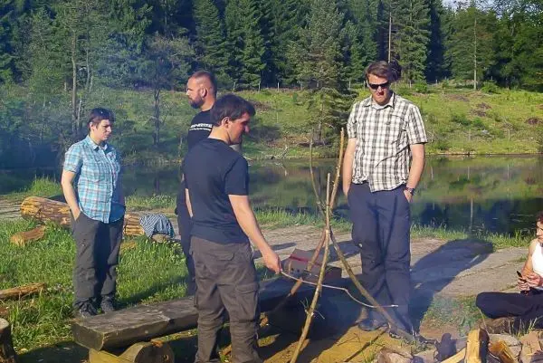 Gruppe steht um ein kleines Bodenfeuer und beobachtet eine improvisierte Konstruktion