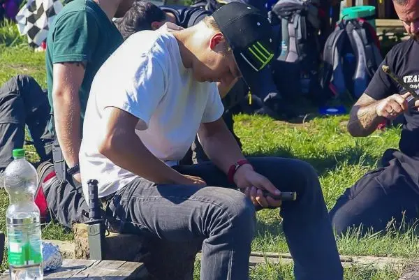 Junger Mann sitzt auf einem Baumstamm und bearbeitet ein Messer