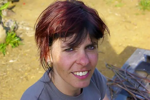 Lächelnde Person mit kurzem, rotem Haar sitzt auf dem Boden neben Holzstücken