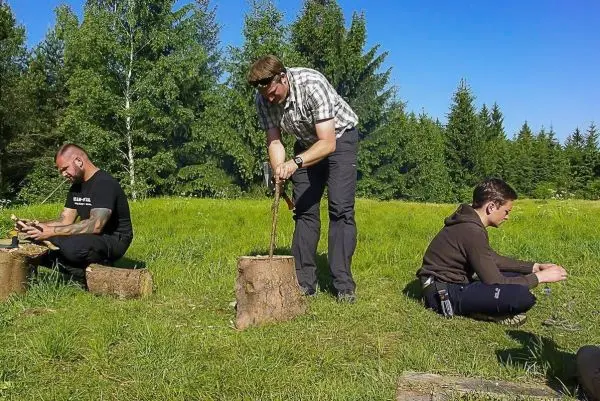 Mann bearbeitet Holzstück mit einem Messer, während zwei weitere Personen auf Baumstümpfen sitzen