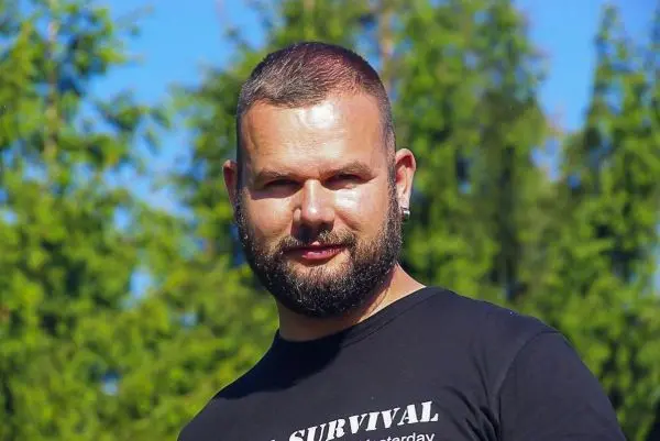 Mann mit Bart trägt schwarzes T-Shirt vor grünen Bäumen