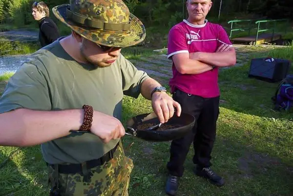 Mann mit Feldmütze bereitet Essen in einer Pfanne vor, während ein anderer zuschaut