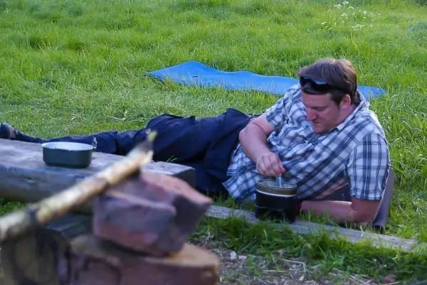 Mann rührt in einem Topf auf einer Holzbank neben einem kleinen Feuer