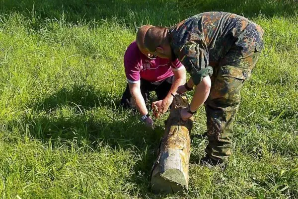 Zwei Personen bearbeiten einen Holzstamm auf einer Wiese