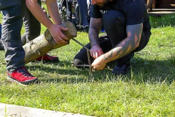 Zwei Personen bearbeiten einen Holzstock mit einem Messer auf dem Gras
