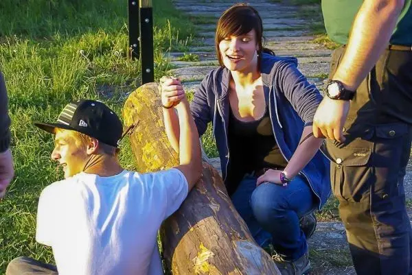 Zwei Personen halten sich an einem Holzstamm fest, während eine dritte Person zuschaut