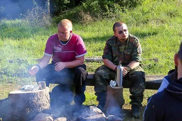 Zwei Personen sitzen an einem Lagerfeuer und halten Tassen in der Hand