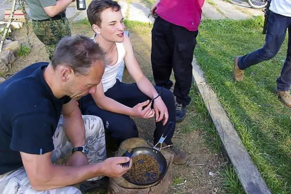 Zwei Personen sitzen neben einem Topf mit Insekten auf einem Holzstück