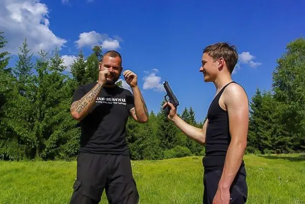 Zwei Personen stehen in einer Wiese, eine hält eine Pistole, die andere hat die Hände an den Kopf