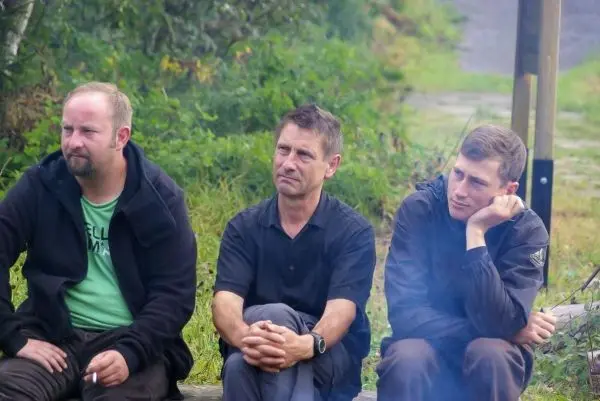 Drei Personen sitzen auf einem Holzstamm und beobachten ein kleines Feuer