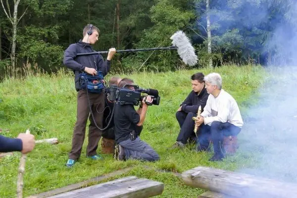 Drei Personen sitzen auf Holzstämmen und diskutieren, während ein Kamerateam filmt
