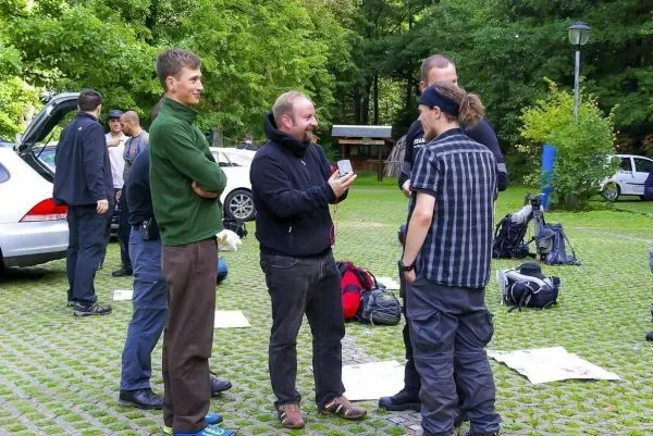 Gruppe diskutiert im Freien, Rucksäcke und Autos im Hintergrund sichtbar