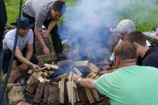 Kleines Bodenfeuer mit Holzscheiten, Rauch steigt auf, mehrere Personen um das Feuer