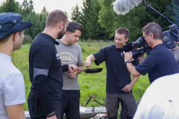 Mann hält Pfanne über Feuerstelle, während andere zuschauen und filmen