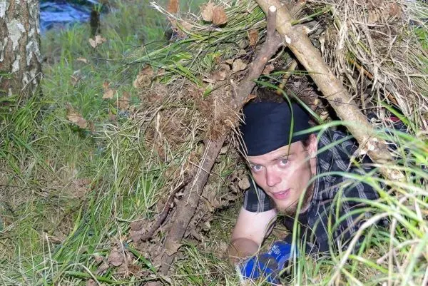 Mann kriecht in eine selbstgebaute Laubhütte aus Ästen und Gras