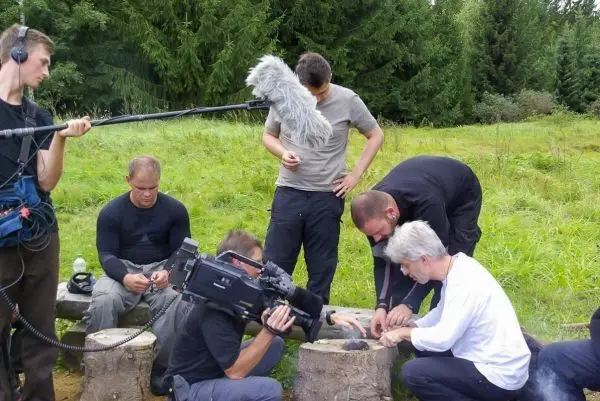 Mehrere Personen arbeiten an einem Holzstück auf Baumstümpfen, Kamera und Mikrofon sind sichtbar