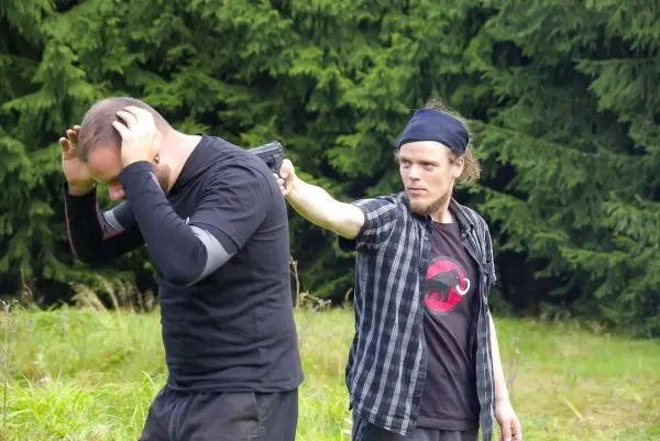 Zwei Personen in einem Wald, eine hält eine Waffe an den Kopf der anderen