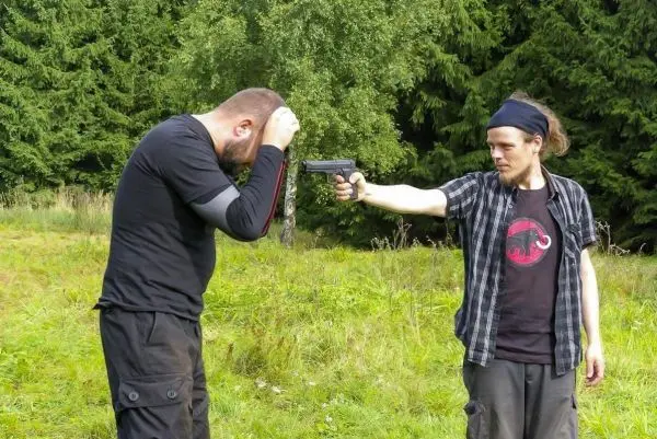 Zwei Personen in einer Wiese, eine hält eine Pistole auf die andere