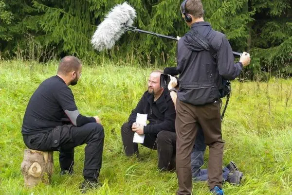 Zwei Personen sitzen auf einem Baumstumpf und diskutieren, während ein Kamerateam filmt