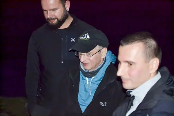 Drei Personen stehen in der Dunkelheit, eine trägt eine Mütze und einen blauen Pullover