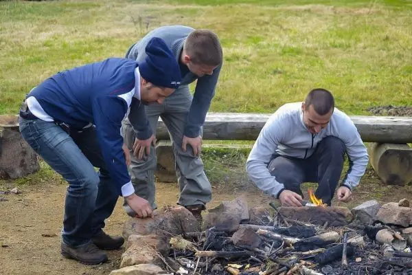 Kleine Flamme brennt in einem Steinkreis, während drei Personen sich darum versammeln