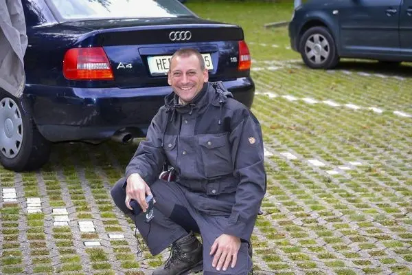 Mann in schwarzer Kleidung kniet neben einem blauen Auto auf Pflastersteinen
