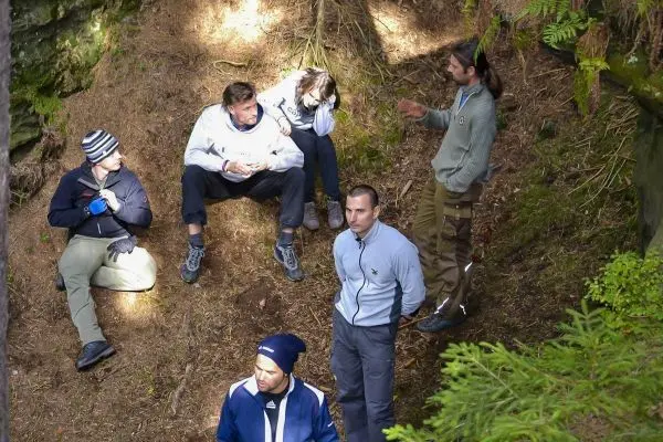 Sechs Personen sitzen und stehen im Wald, einige diskutieren miteinander