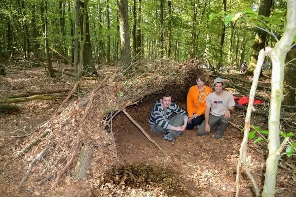 Drei Personen sitzen vor einer selbstgebauten Tarp-Notunterkunft im Wald