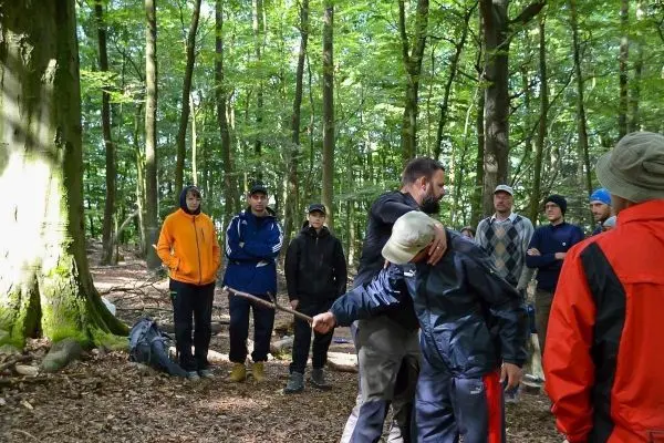 Ein Mann demonstriert eine Technik mit einem Stock, während Zuschauer im Wald beobachten