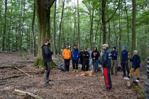 Gruppe hört einem Vortrag im Wald, im Hintergrund liegt ein Hund