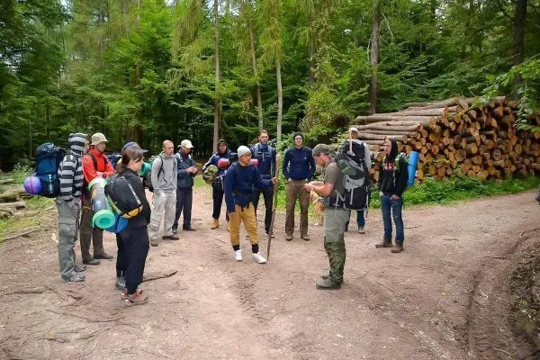 Gruppe mit Rucksäcken steht an einem Waldweg und hört einem Anleiter zu