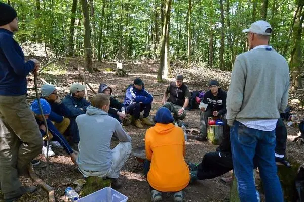 Gruppe sitzt im Wald auf Baumstümpfen und hört einem Vortrag zu