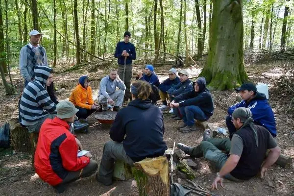 Gruppe sitzt um ein Feuer in einem Wald, Holzstämme als Sitzgelegenheiten