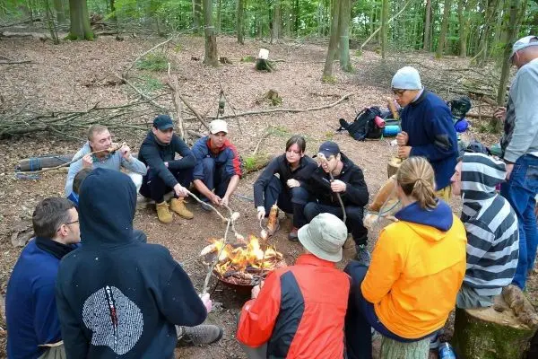 Gruppe sitzt um ein kleines Feuer und röstet Essen auf Stockspießen