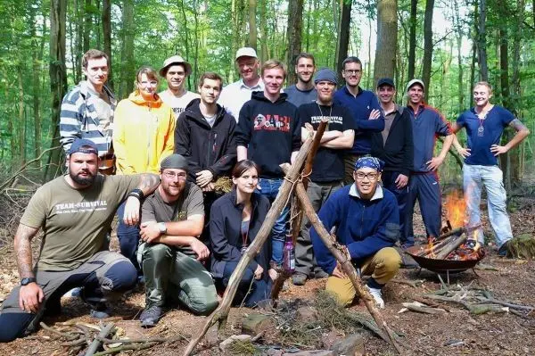 Gruppierung von Personen vor einem Lagerfeuer und einer Holzstruktur im Wald