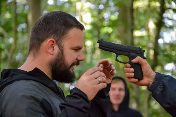 Hand hält eine Pistole in Richtung eines Mannes mit Bart im Wald