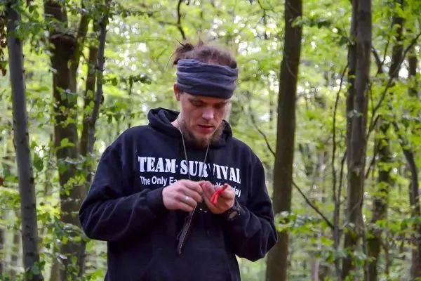 Mann mit Stirnband hält ein rotes Objekt in der Hand, umgeben von Bäumen