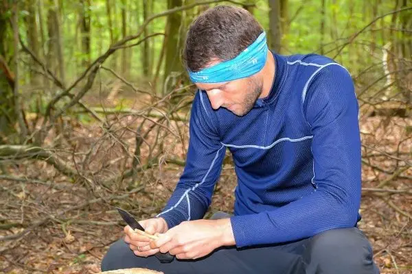 Mann sitzt im Wald und bearbeitet Holzstück mit einem Messer