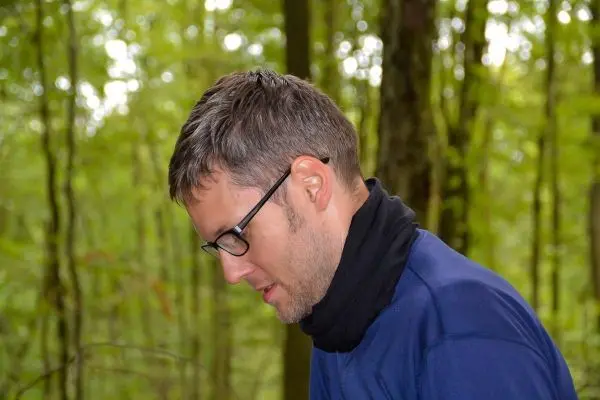 Person mit Brille und Schal beugt sich in einem Wald nach unten